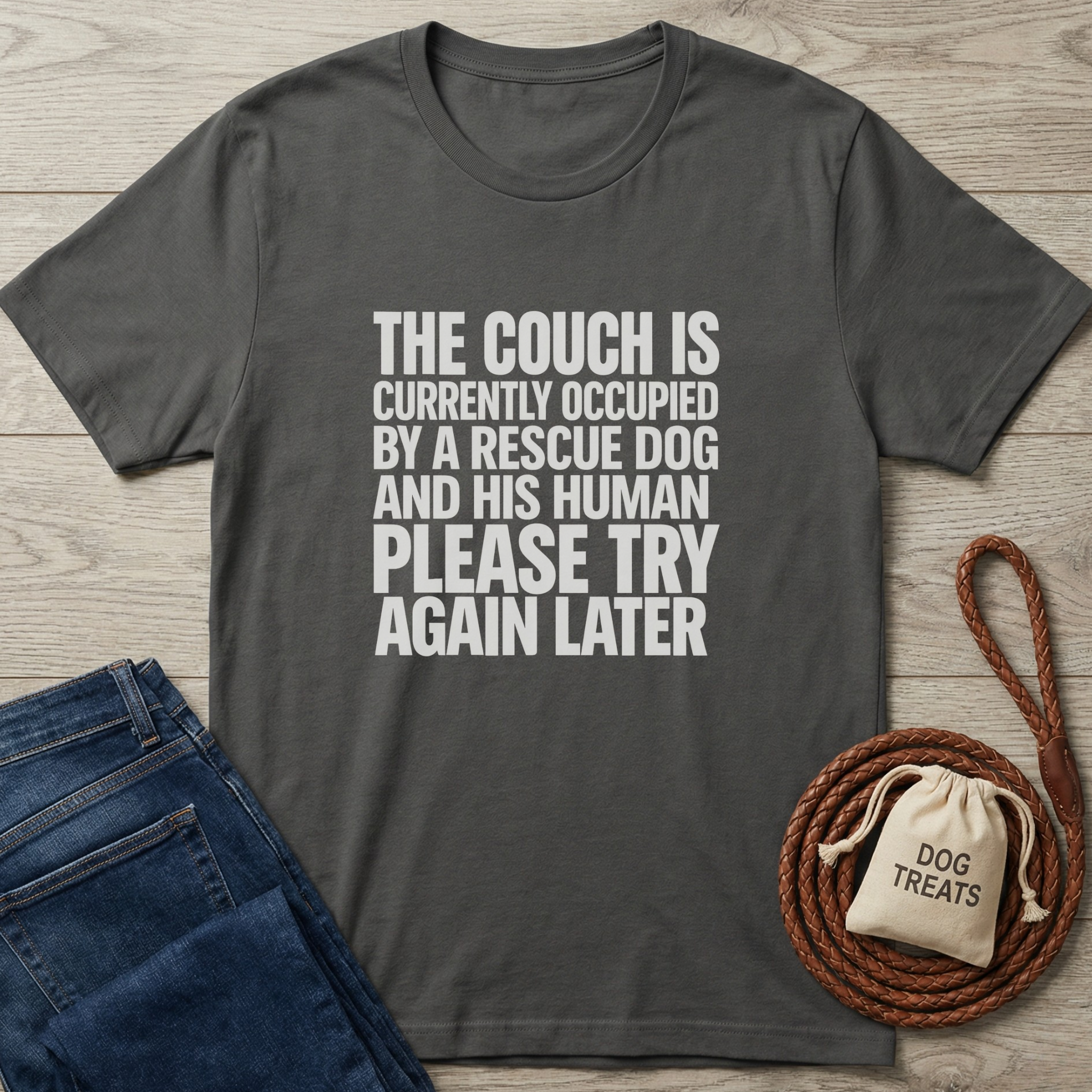 Charcoal gray cotton t-shirt with "THE COUCH IS CURRENTLY OCCUPIED BY A RESCUE DOG..." white text, next to jeans and dog treats bag.