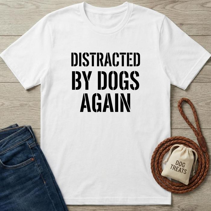 White t-shirt with 'Distracted by Dogs Again' text, next to jeans and dog-related items on a wooden surface.