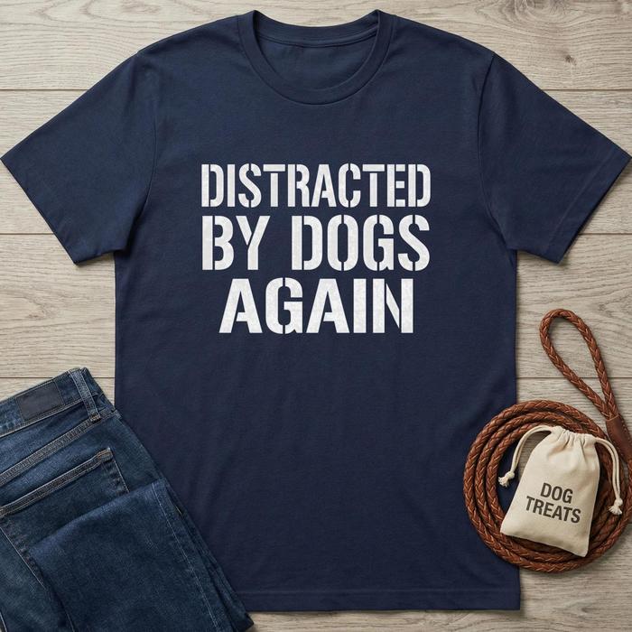 Navy blue t-shirt with 'DISTRACTED BY DOGS AGAIN' text on a wooden surface with jeans and dog treats bag.