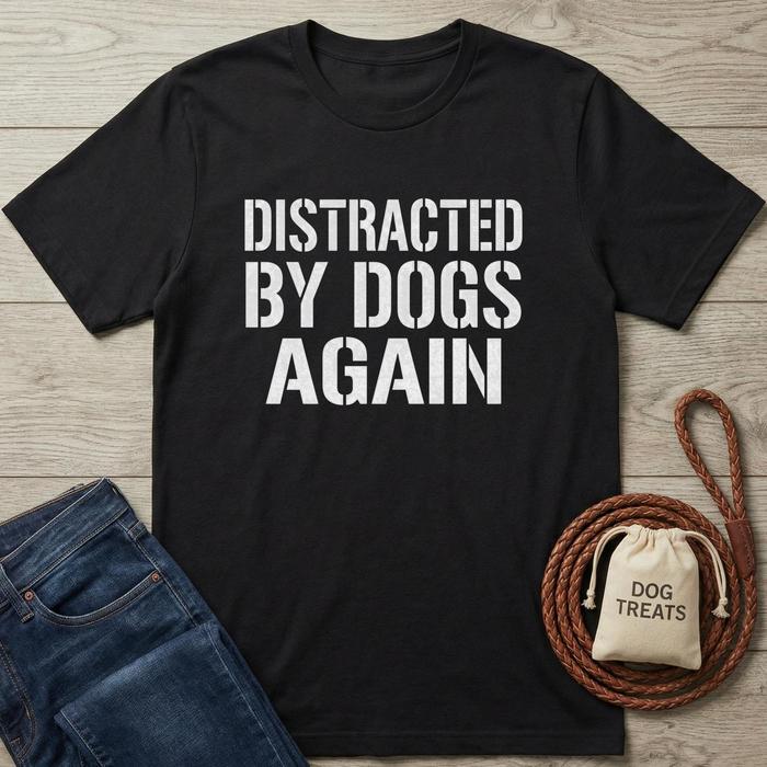 Black t-shirt with 'DISTRACTED BY DOGS AGAIN' text, next to jeans and a dog treat bag on a wooden surface.