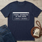 Navy blue t-shirt with 'Status: Distracted by Dogs Again' text on a wooden surface with jeans and a dog treat bag.
