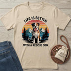 Beige t-shirt with a graphic of a dog and text 'Life is better with a rescue dog' on a wooden surface with jeans and a 'Dog Treats' bag.