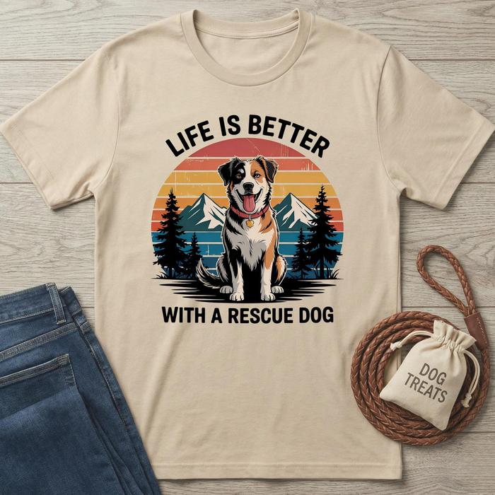 Beige t-shirt with a graphic of a dog and text 'Life is better with a rescue dog' on a wooden surface with jeans and a 'Dog Treats' bag.