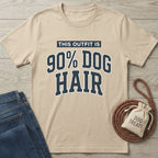 Beige cotton t-shirt with "THIS OUTFIT IS 90% DOG HAIR" in navy varsity font, next to jeans and dog treats.