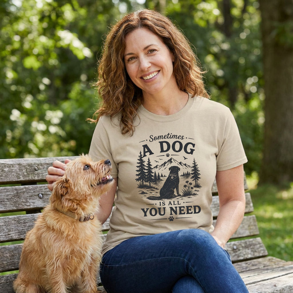 Woman in a tan "Sometimes a Dog Is All You Need" graphic tee petting a small brown dog on a park bench.