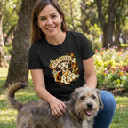 Woman in a black "Rescued & Rad" t-shirt with a smiling dog design, petting a scruffy brown dog outdoors.