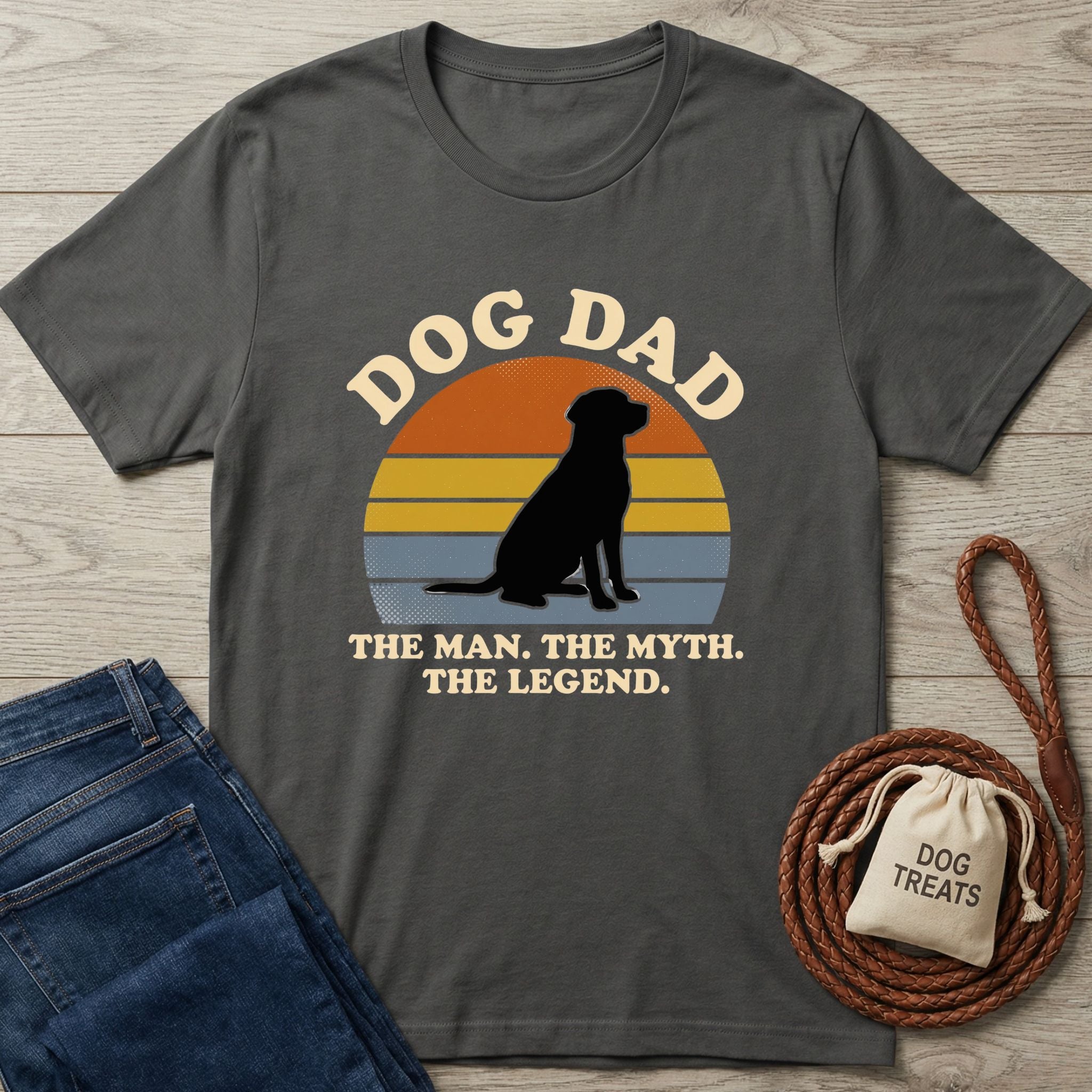 Charcoal gray "Dog Dad" t-shirt with a black dog silhouette against a sunset graphic, next to jeans and dog treats.