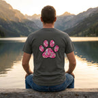 Man in a charcoal Gildan Softstyle tee with a large pink floral paw print design on the back, sitting by a lake.