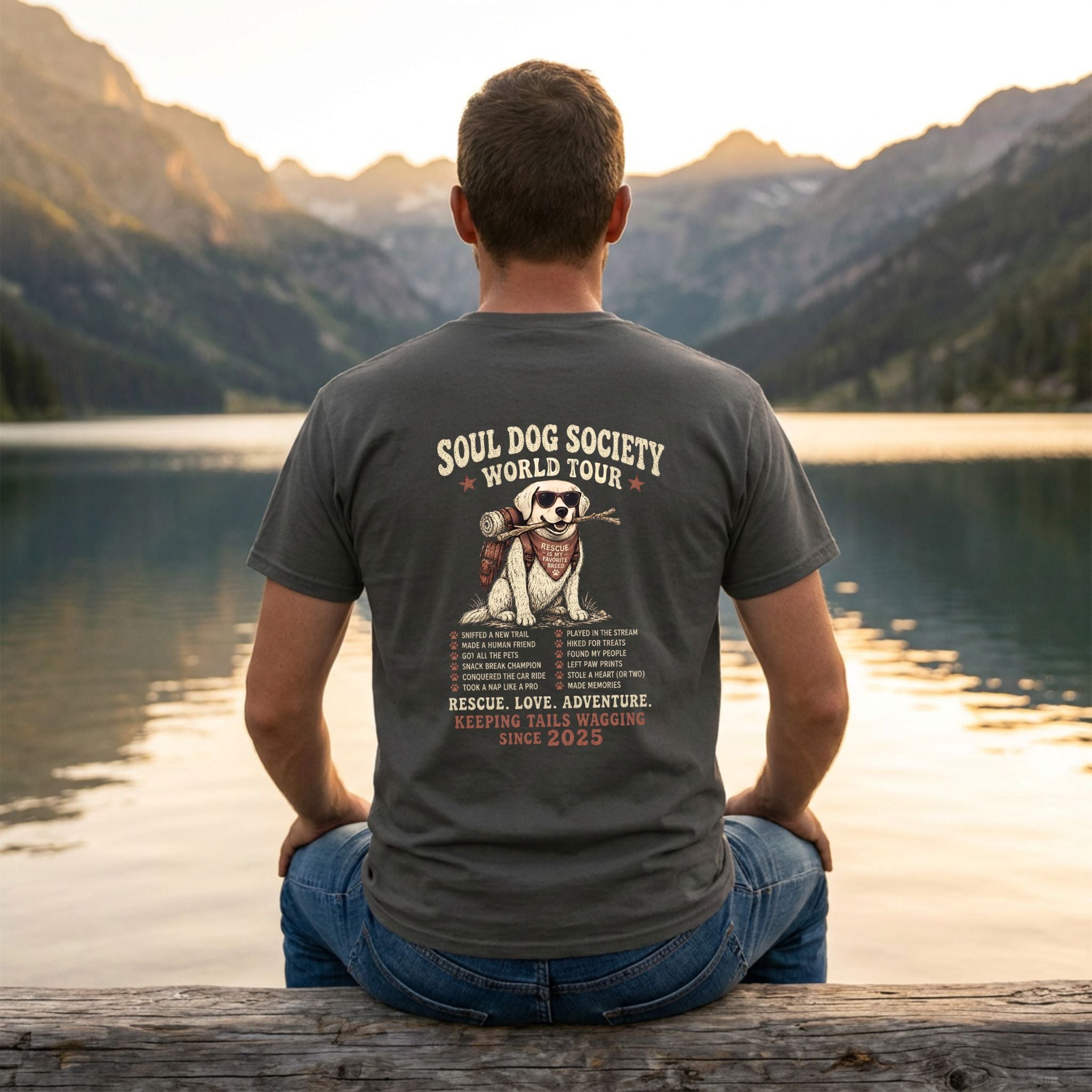 Man in a charcoal grey Soul Dog World Tour T-shirt with a graphic of a dog, sitting by a lake at sunset.