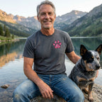 Man in a charcoal grey Paws For Hope T-Shirt with a pink floral paw print, sitting by a lake with a dog.
