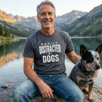 Man in a charcoal grey "Easily Distracted By Dogs" t-shirt and jeans, sitting with a blue heeler dog by a mountain lake.