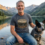 Man in a charcoal gray Silver Muzzle Society t-shirt with a sleeping dog design, sitting by a lake with his blue heeler.