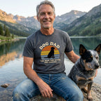 Smiling man in a charcoal "Dog Dad" tee with a sunset and dog silhouette, next to a blue heeler by a mountain lake.