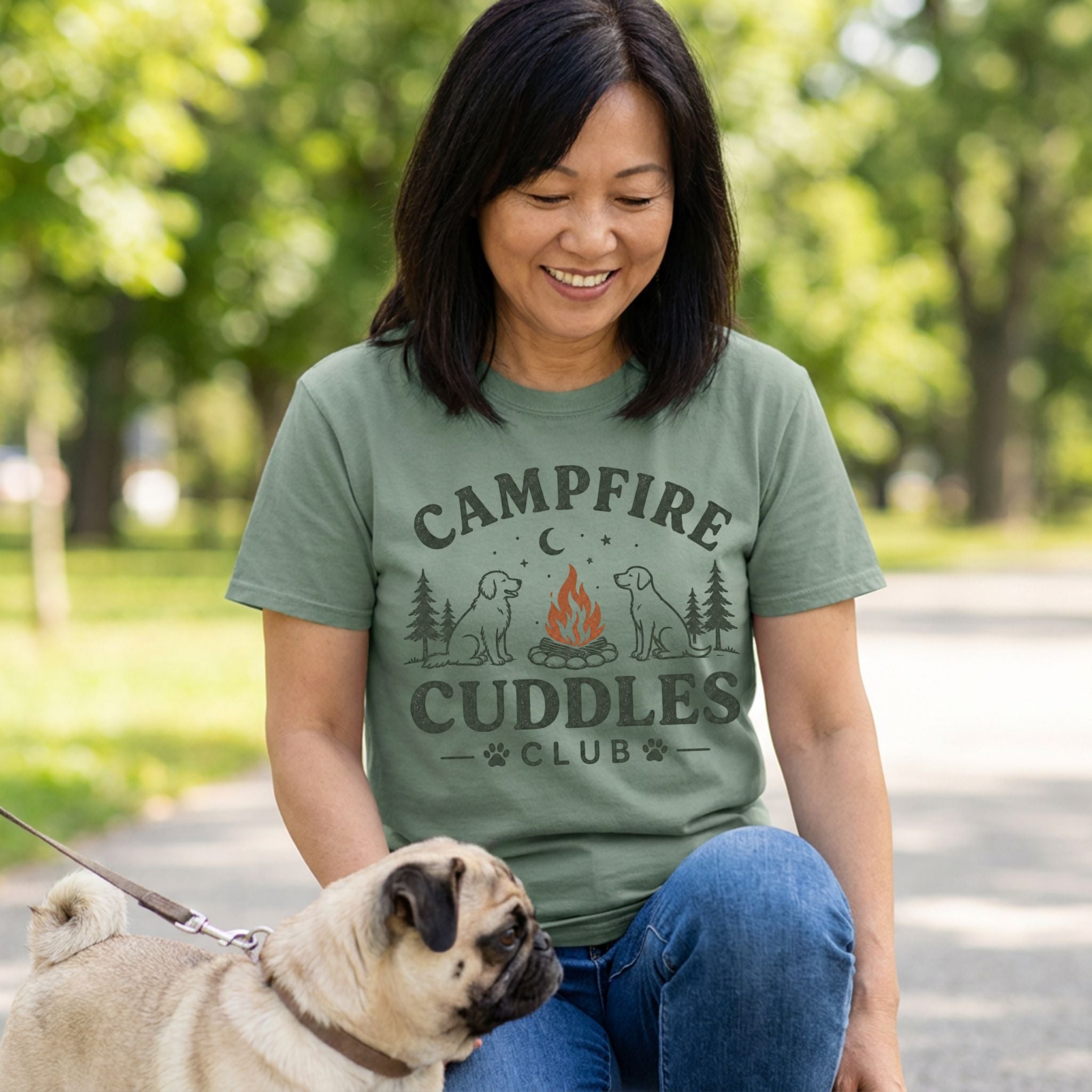 Woman wearing a sage green crewneck t-shirt with a campfire and dog graphic, kneeling outdoors with her pug.