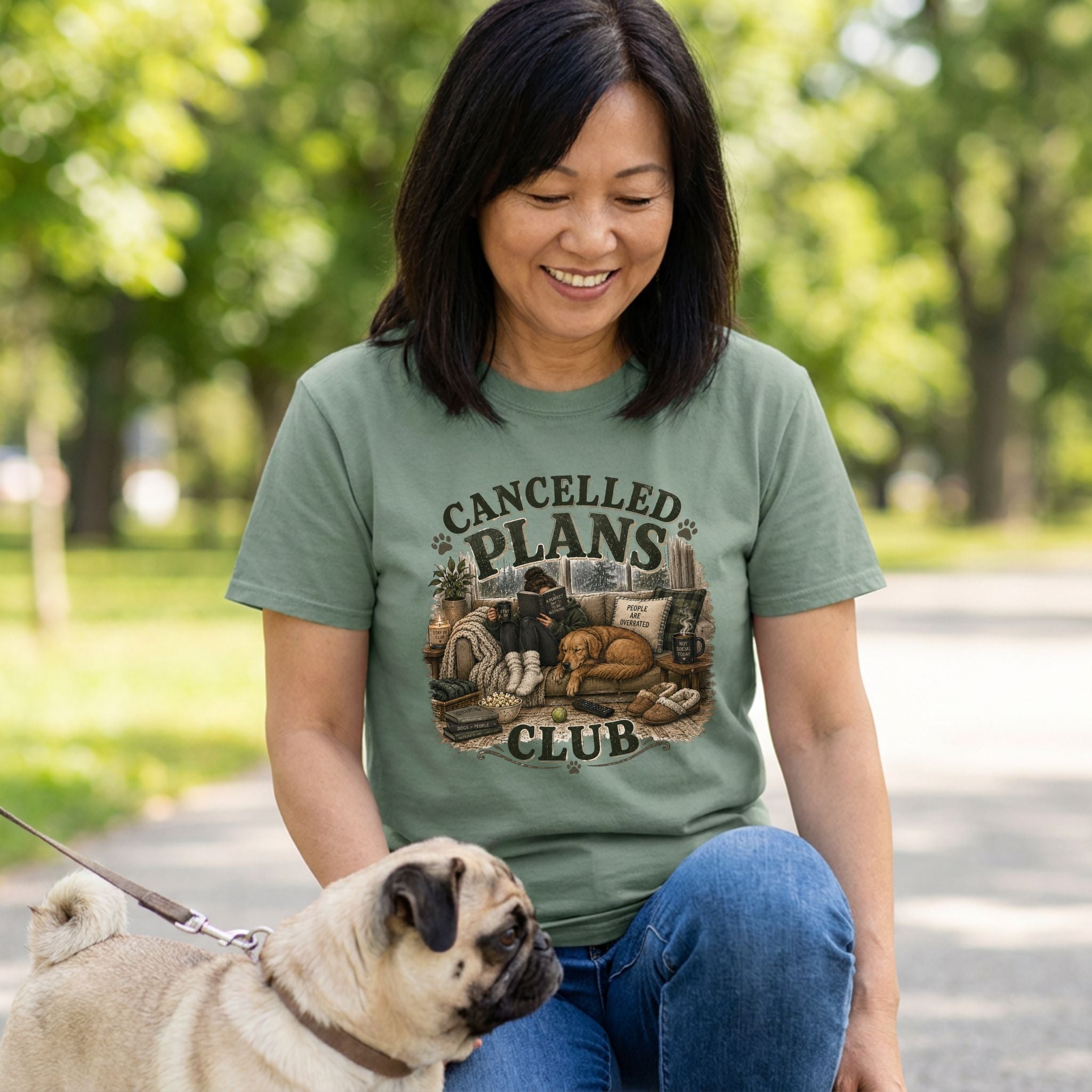 Woman wearing a sage green crewneck t-shirt featuring a graphic of a person reading on a couch with a dog.