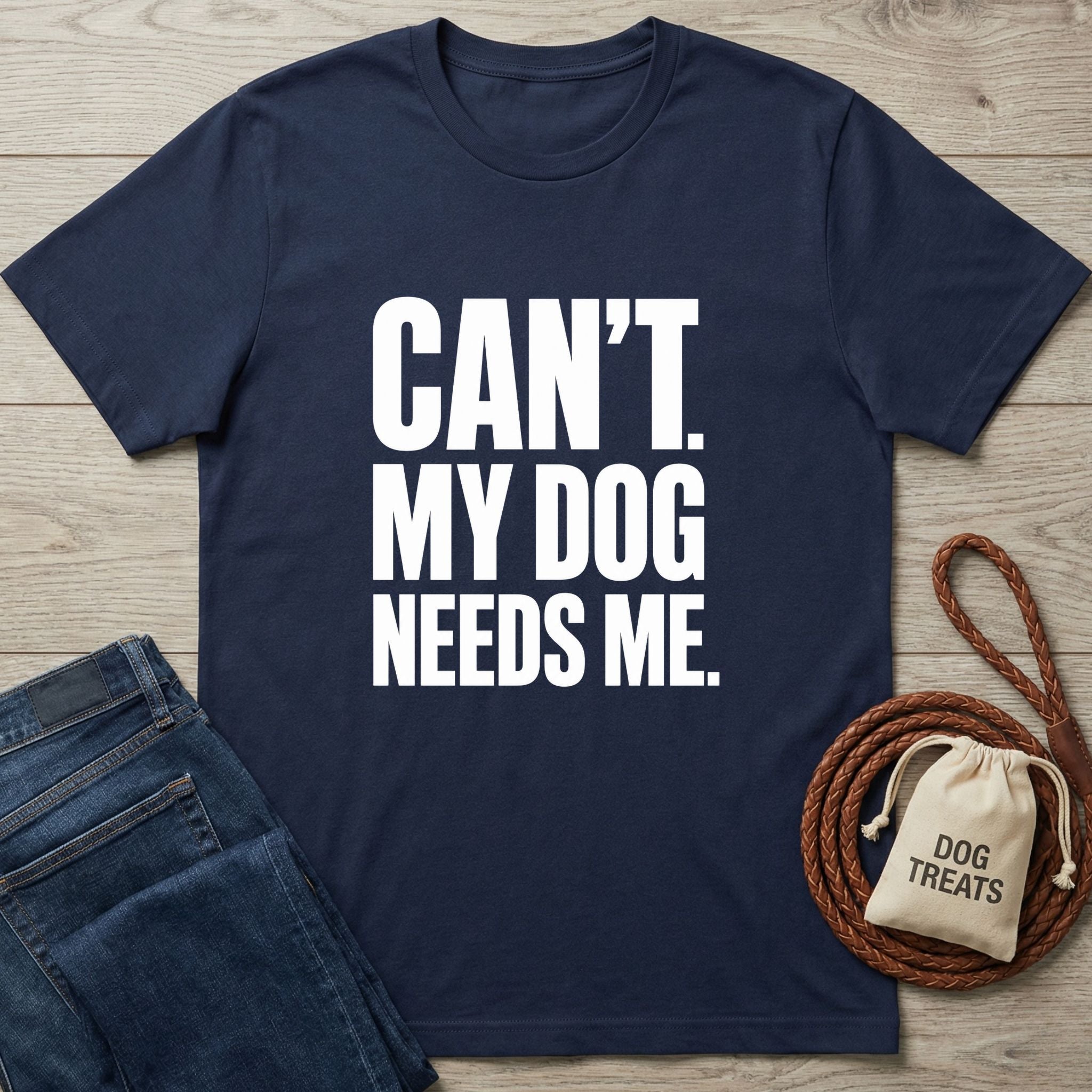 Navy blue crew neck t-shirt with white text reading CAN'T. MY DOG NEEDS ME, laid on wood with jeans and a dog treat bag.