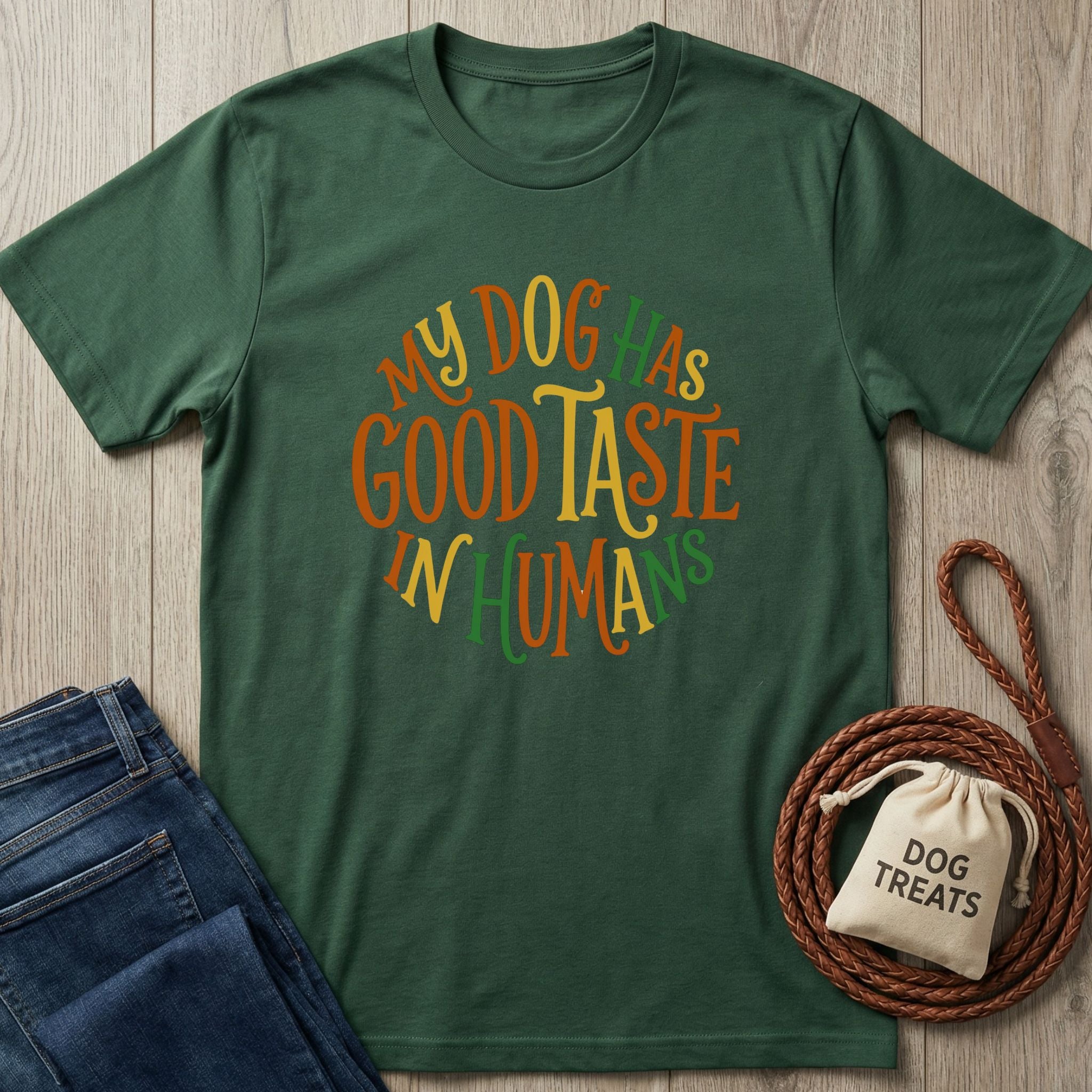 Forest green Gildan Softstyle® t-shirt with "My Dog Has Good Taste In Humans" text, next to jeans and dog treats.