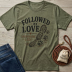 Olive green "Followed By Love Everywhere I Go" t-shirt with a boot print and paw prints, next to jeans and dog treats.