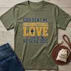 Olive green "God Sent Me Unconditional Love In A Rescue Dog" tee with blue and gold distressed text, beside jeans and dog accessories.