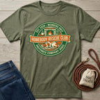 Green t-shirt with 'Homebody Rescue Club' design on a wooden surface with jeans and dog treats bag.