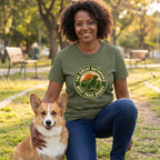 Woman in an olive green "Best Trail Buddy" t-shirt with a dog graphic, kneeling beside a corgi outdoors.