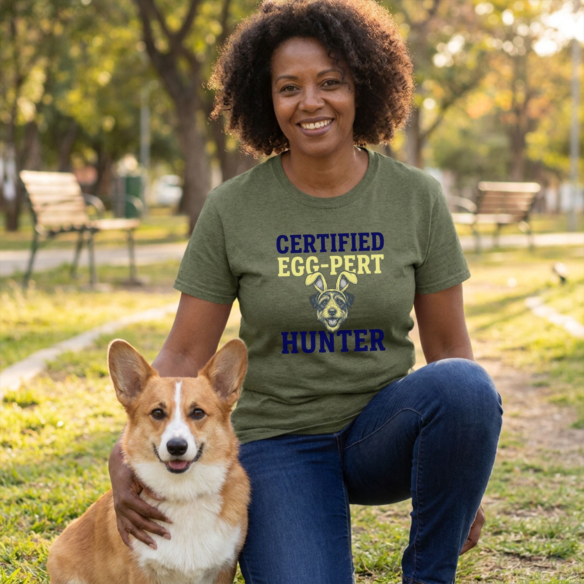 Woman in olive green "Certified Egg-Pert Hunter" t-shirt with a dog graphic, kneeling with a corgi outdoors.