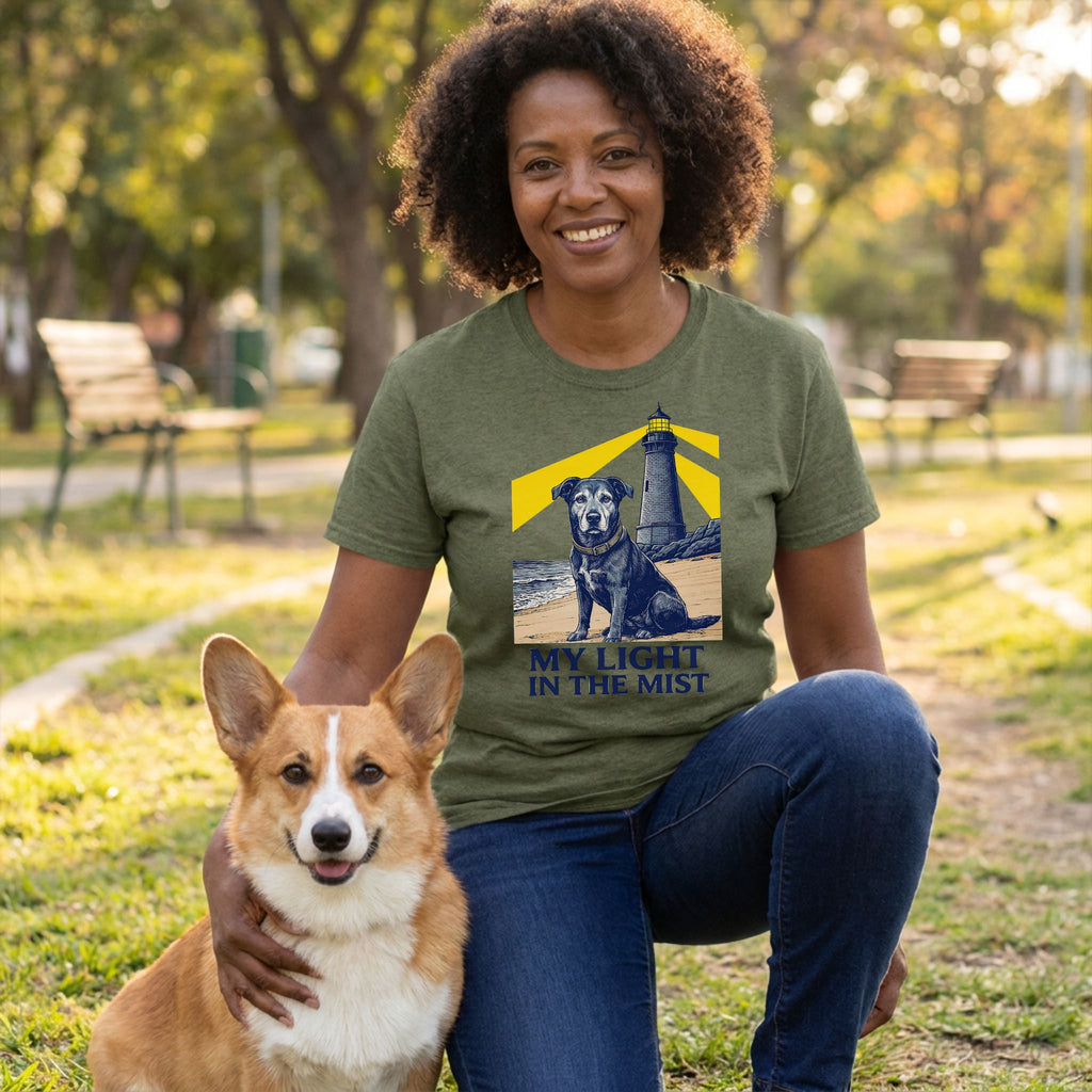 Woman in olive green "My Light In The Mist" t-shirt with rescue dog and lighthouse graphic, kneeling with a corgi.