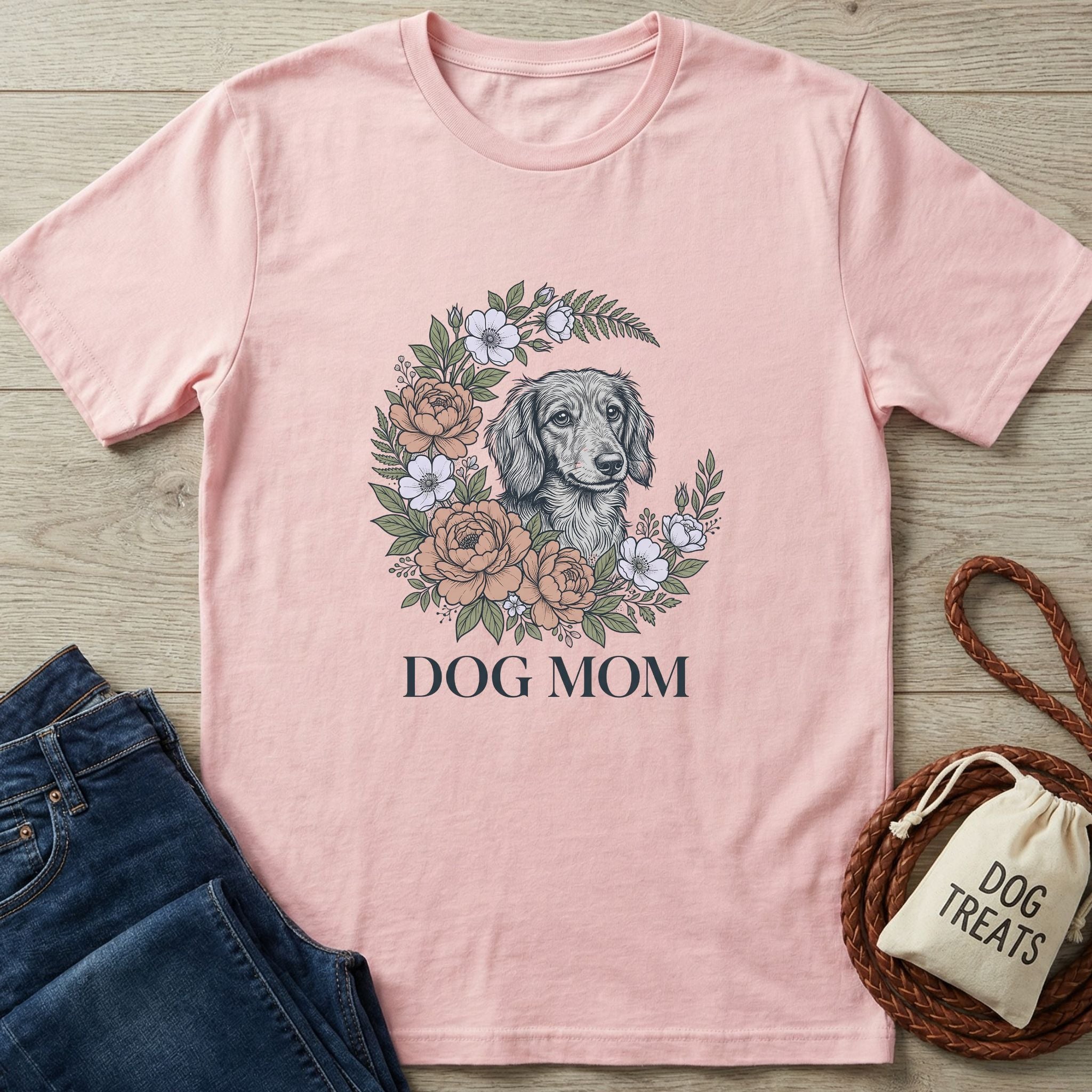 Pink Gildan Softstyle Dog Mom t-shirt with a floral wreath and dachshund graphic, next to jeans and dog treats.