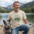 Man in a beige "Lead Me To The Rock" graphic tee with a rescue dog by a mountain lake.