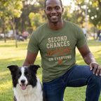 Man wearing an olive green crewneck t-shirt with Second Chances Look Good on Everyone text, kneeling beside a dog.