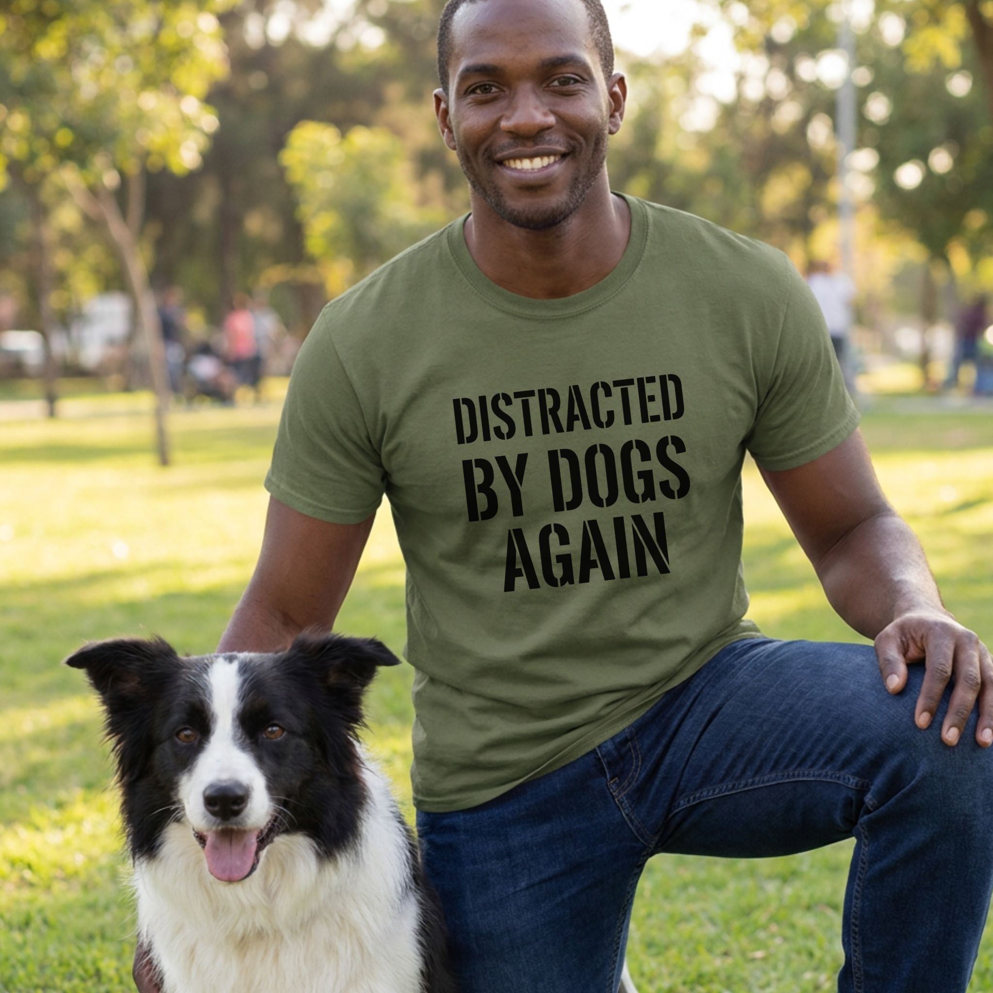 Man in an olive green "Distracted By Dogs Again" t-shirt kneels beside a black and white Border Collie.