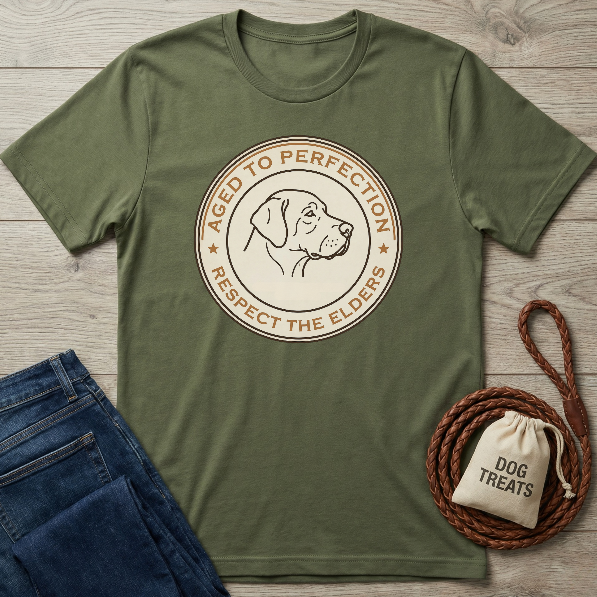 Olive green "Aged To Perfection" t-shirt with a dog profile graphic, next to folded jeans and dog treats.