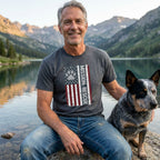 Man in a charcoal grey "Mission: Rescue" tee with an American flag and paw print design, sitting with a dog by a lake.