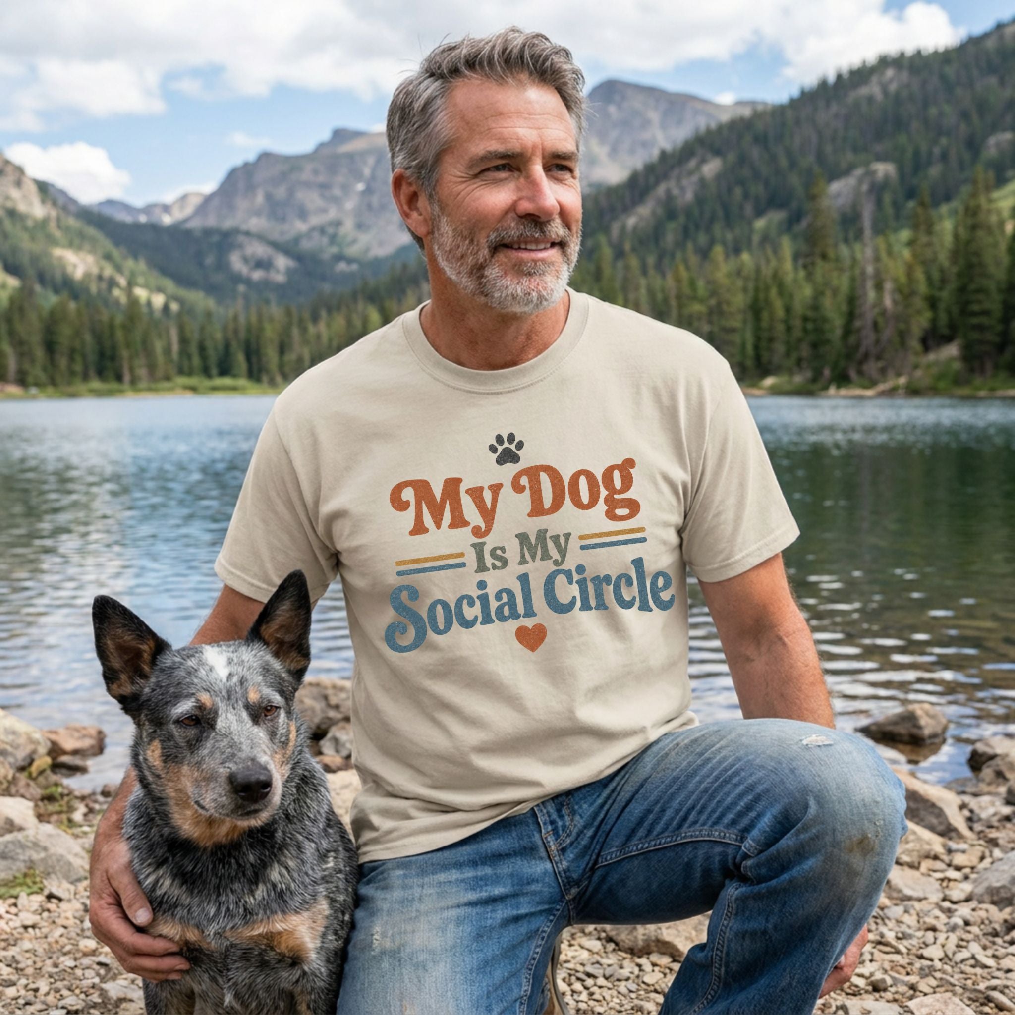 Man wearing a beige crewneck t-shirt with My Dog Is My Social Circle text, sitting by a lake with his cattle dog.