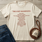 Cream t-shirt with "The Cozy Manifesto" text in brown, paired with jeans and a dog treats bag.