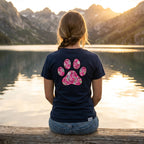 Woman in a navy Gildan Softstyle tee with a pink ribbon paw print design, sitting by a lake at sunset.