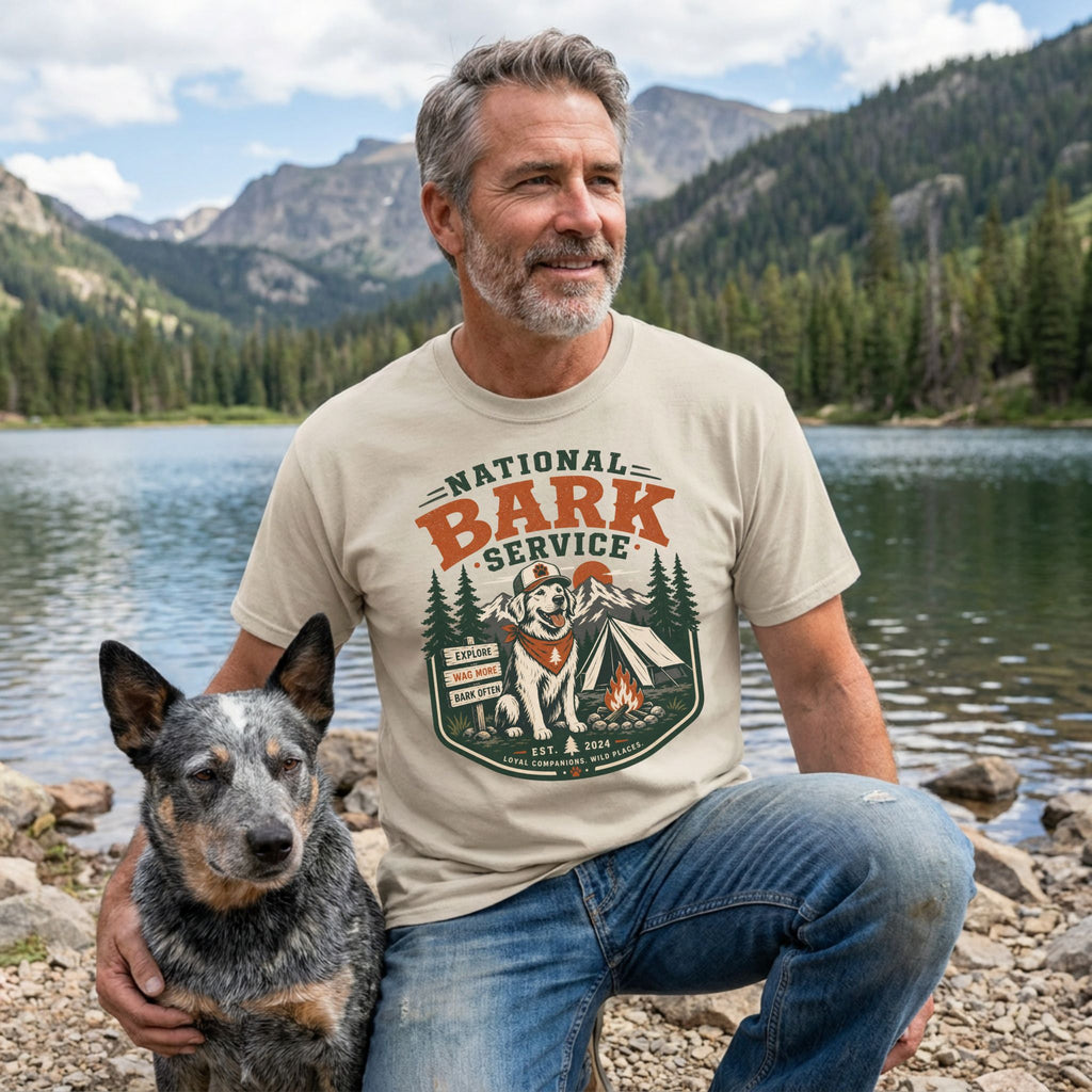 Man wearing a cream-colored National Bark Service graphic t-shirt featuring a dog, tent, and mountain scene by a lake.