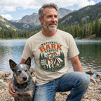 Man wearing a cream-colored National Bark Service graphic t-shirt featuring a dog, tent, and mountain scene by a lake.