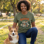 Woman wearing an olive green National Bark Service graphic t-shirt while kneeling next to a corgi in a park.