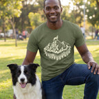 Man in an olive green "King of The Hill" t-shirt with a mountain and dog graphic, kneeling beside a Border Collie.