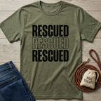 Olive green "Rescued" t-shirt, jeans, braided leather leash, and dog treats bag on wood.