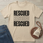 Tan "Rescued" graphic tee with blue jeans, braided leash, and "Dog Treats" bag on a wooden surface.