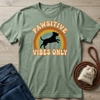 Sage green Pawsitive Vibes Only T-shirt with a jumping dog and rainbow graphic, next to jeans and dog treats.