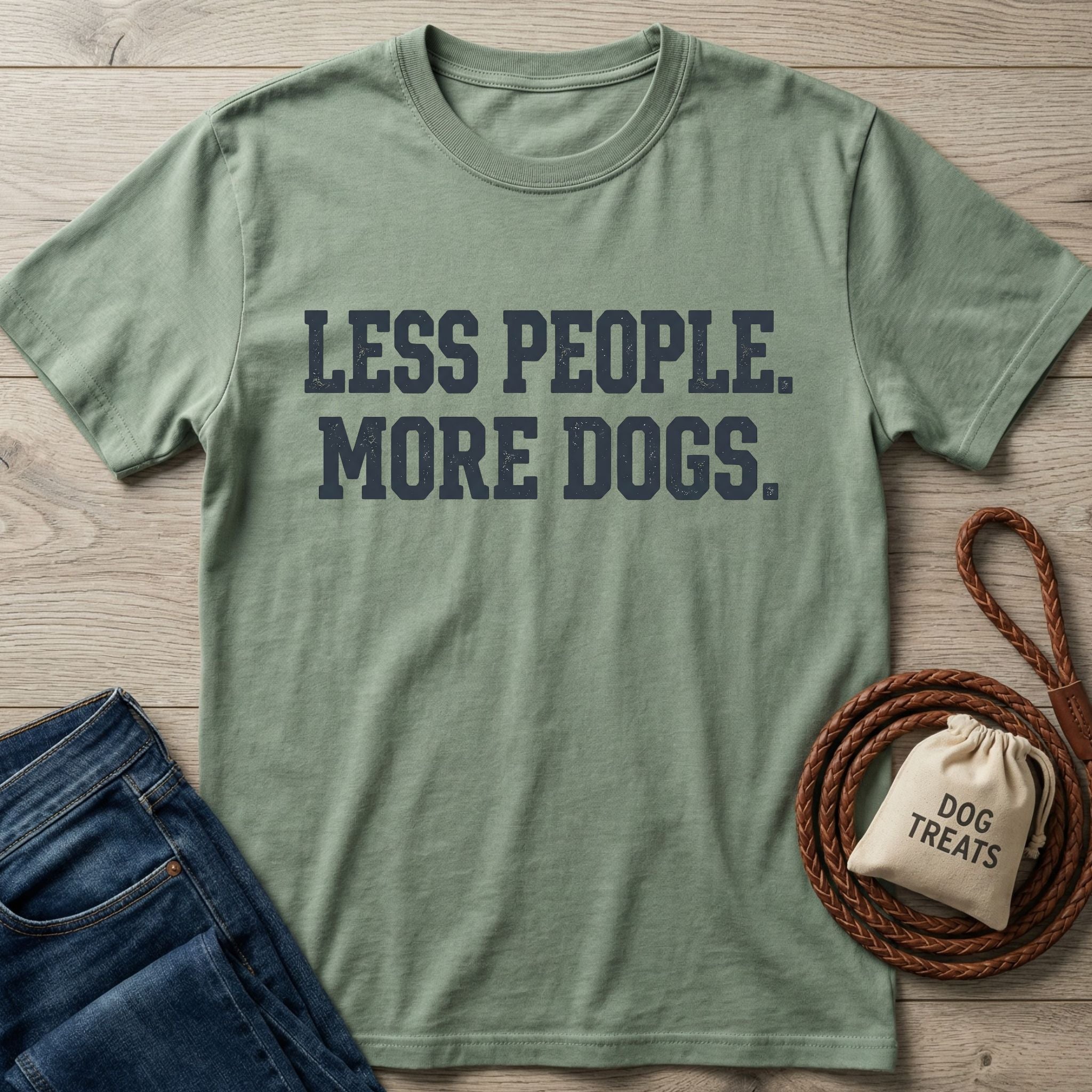 Sage green "Less People. More Dogs." t-shirt with jeans, dog leash, and treat bag on wood.