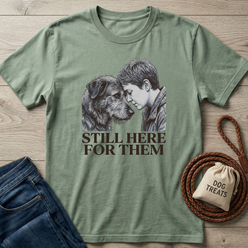 Sage green "Still Here For Them" t-shirt with a graphic of a person and a dog, next to jeans and dog treats.