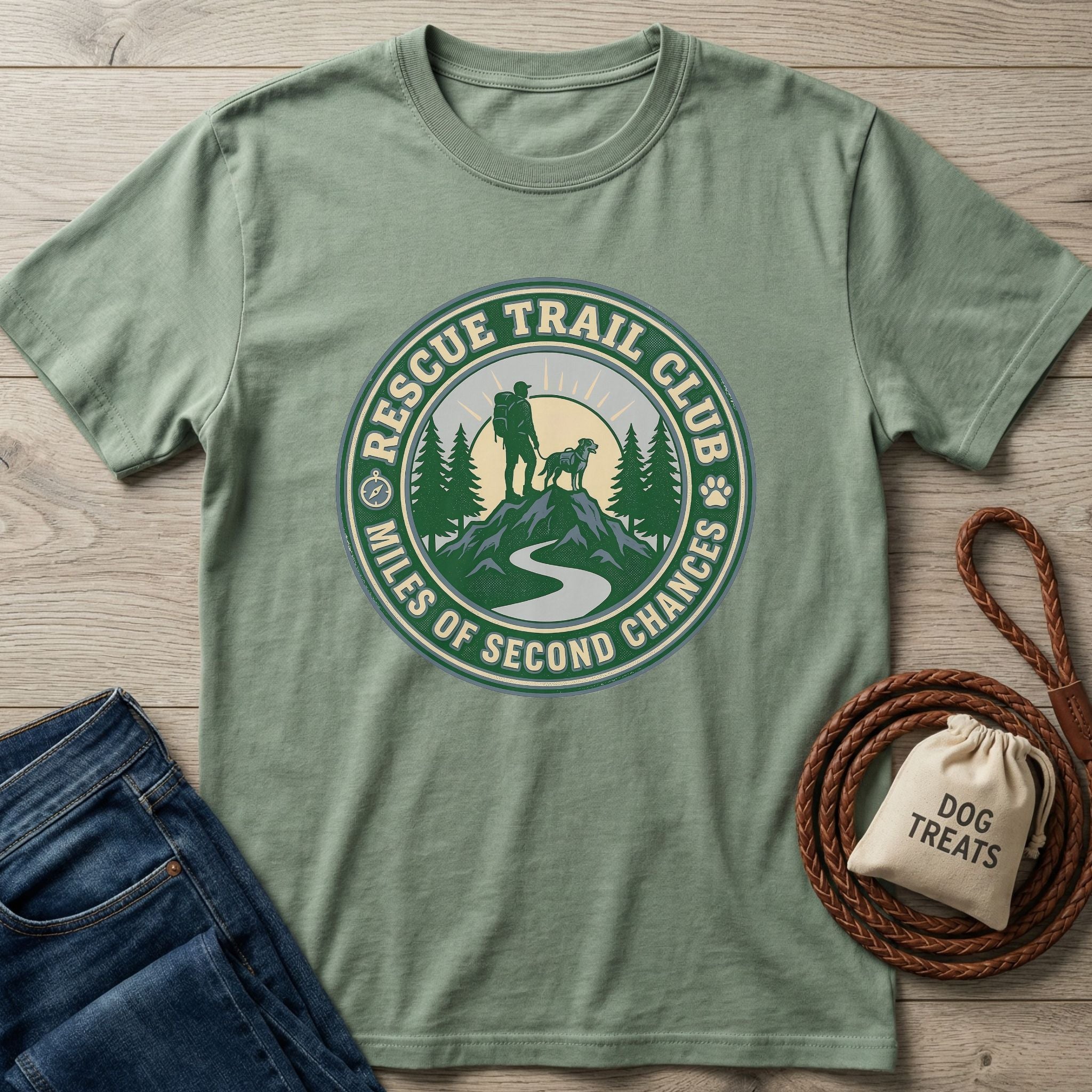 Sage green Rescue Trail Club T-shirt with a graphic of a hiker and dog on a mountain, next to jeans and dog treats.