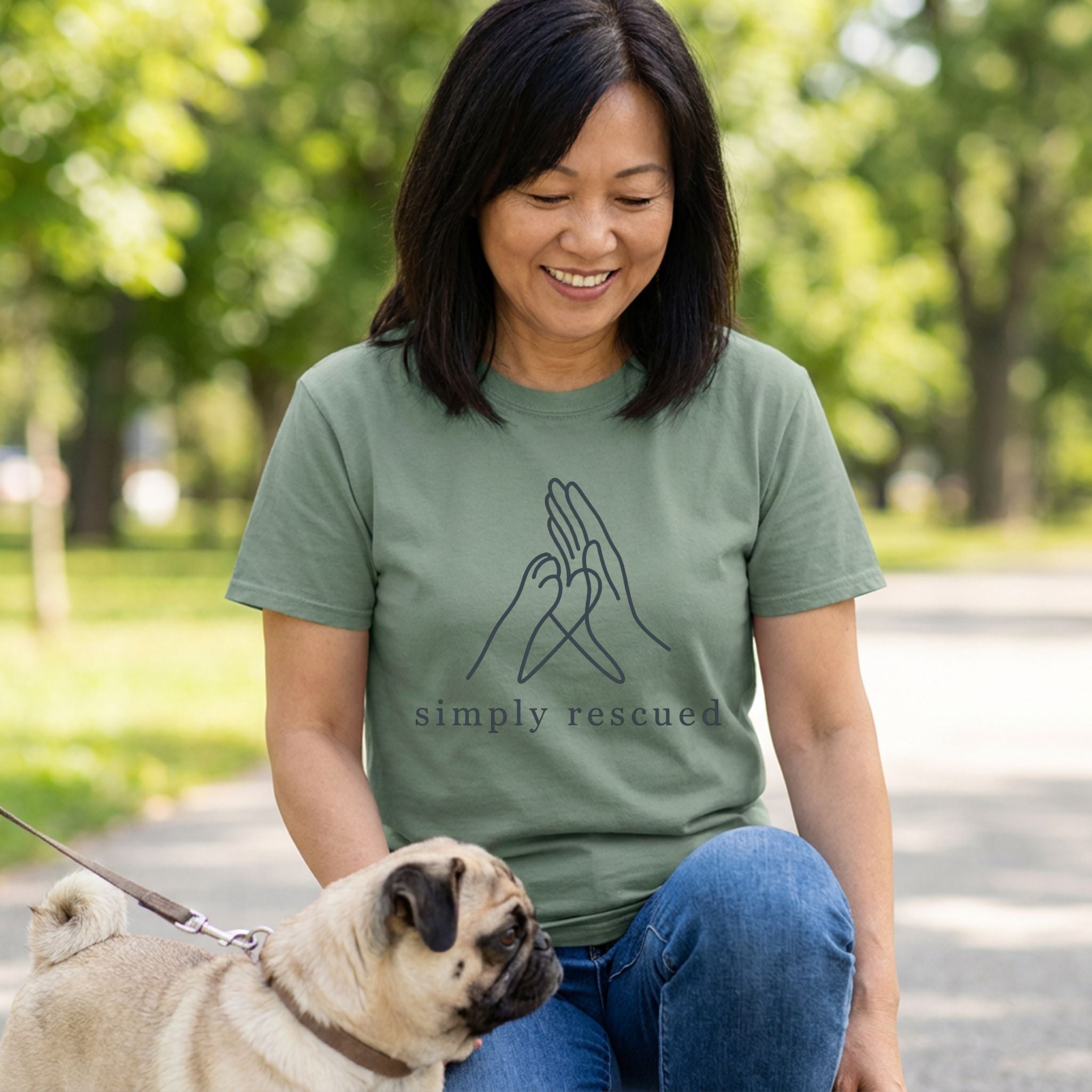 Woman in an olive green "Simply Rescued" tee with a paw and hand design, petting a pug.