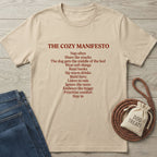 Tan Gildan Softstyle t-shirt with "The Cozy Manifesto" text in red, next to jeans and a dog treats bag.