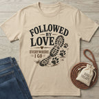 Beige cotton t-shirt with "Followed By Love Everywhere I Go" text, a boot print, and paw prints.