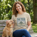Woman in a tan "Trail Tested" graphic tee petting a small brown dog on a park bench.